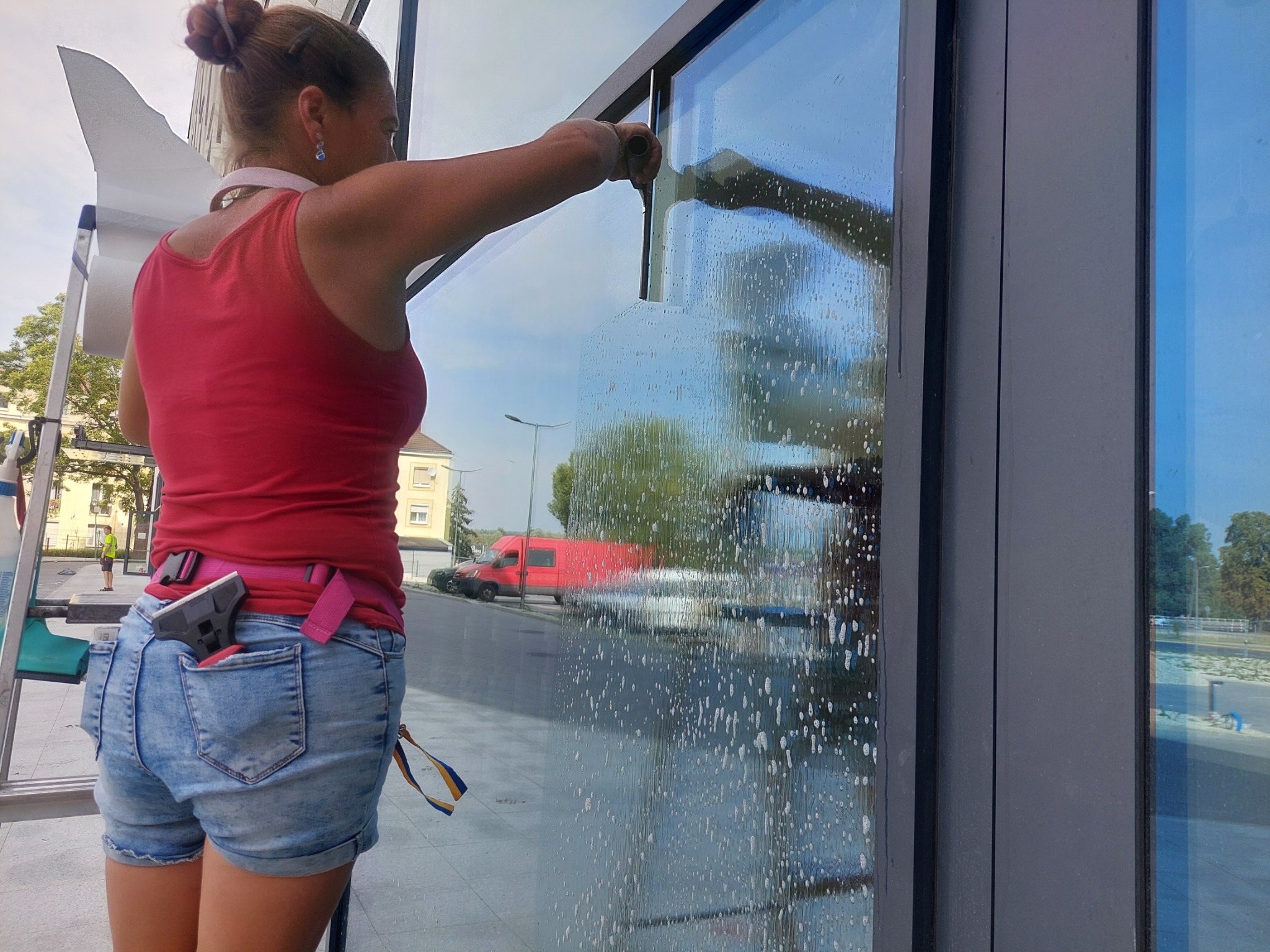 Kobieta w czerwonym topie czyści dużą szybę za pomocą ściągaczki, widoczne smugi wody i piany, w tle czerwony van i budynek.