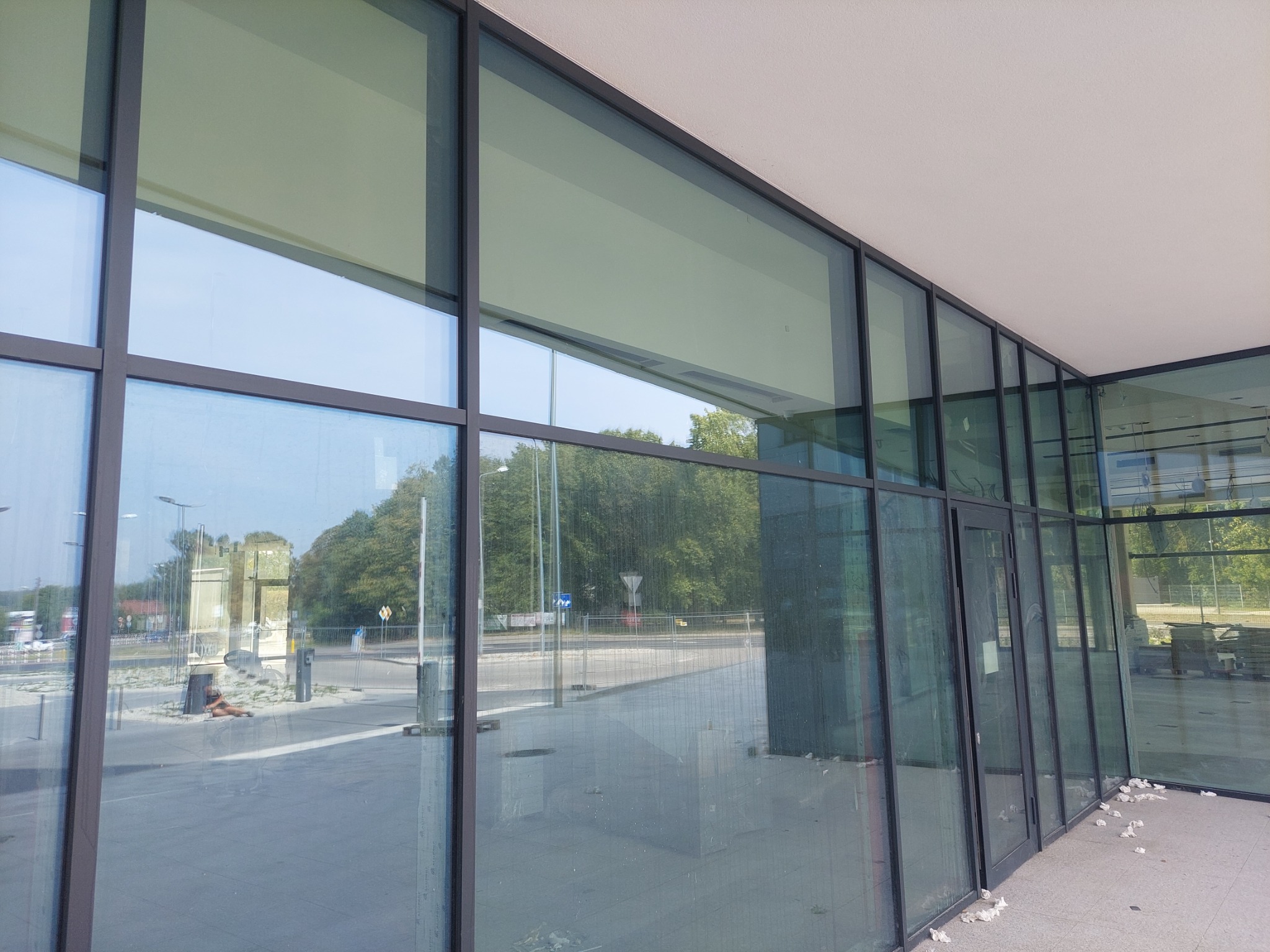 Szklana fasada budynku z widocznymi śladami po remoncie: resztki zaprawy i gruzu na chodniku przed wejściem. Odbicie w szybach ukazuje drzewa i elementy infrastruktury miejskiej.