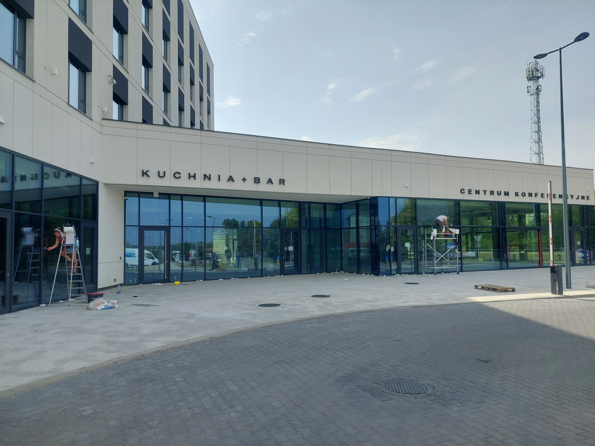 Kobieta na drabinie myje duże okna budynku z napisem 'KUCHNIA+BAR' i 'CENTRUM KONFERENCYJNE', widoczne ślady po remoncie na chodniku.