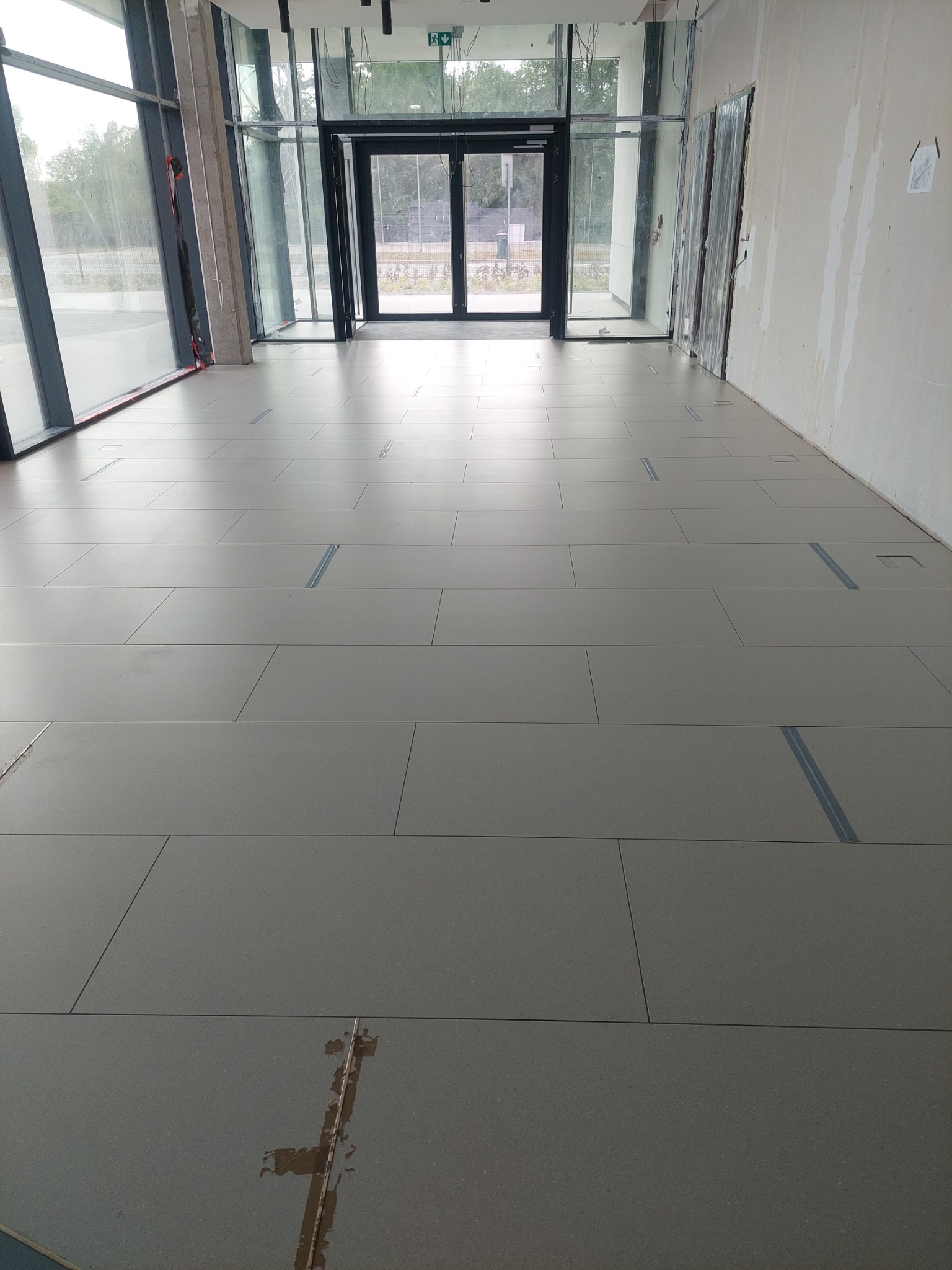 Nowo ułożona szara podłoga z płytek w przestronnym korytarzu, widoczne ślady po remoncie na płytkach w dolnej części kadru, duże okna i szklane drzwi w tle.