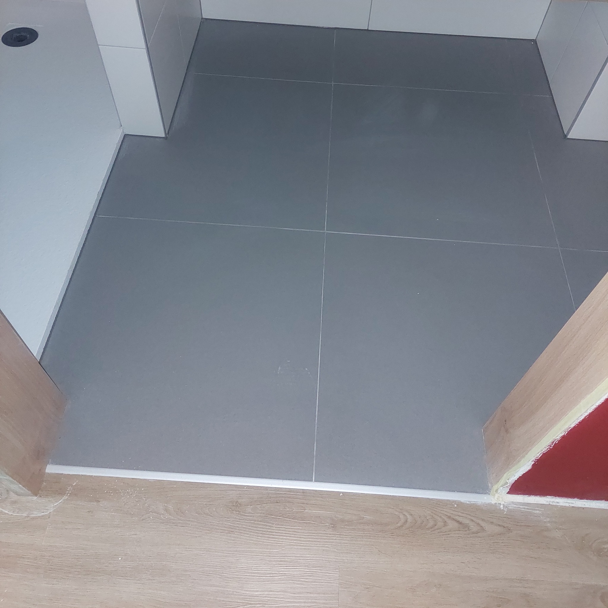 Szare płytki podłogowe ułożone w łazience, widoczne fugi i listwa przypodłogowa, fragment drewnianej podłogi przy wejściu.