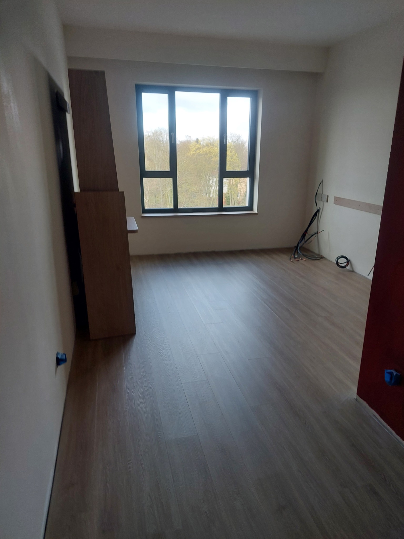 Puste pomieszczenie w Obornikach z panelami podłogowymi i widokiem na drzewa za oknem, przygotowane do prac wykończeniowych.