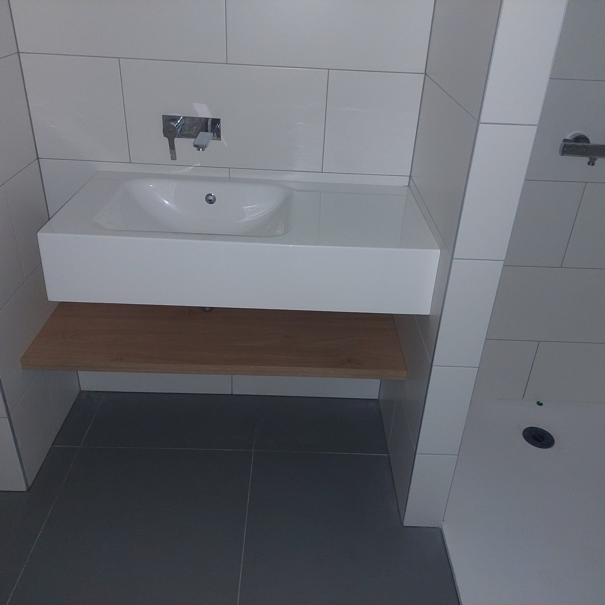 Nowoczesna umywalka z białą szafką i drewnianą półką w minimalistycznej łazience z szarymi płytkami podłogowymi i białymi płytkami ściennymi.