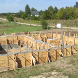 Wylane fundamenty domu jednorodzinnego, drewniane szalunki, widok z góry, teren budowy z widokiem na okoliczne domy i drzewa.