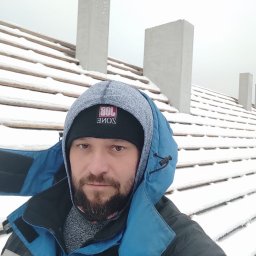 Gedbud Grzegorz Ged - Selfie dekarza na tle zasypanego śniegiem dachu z kominami, ubrany w zimową kurtkę i czapkę z logo.