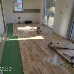 Gedbud Grzegorz Ged - Wnętrze w trakcie układania drewnianej podłogi, widoczne panele, podkład z zielonej pianki, narzędzia i opakowania po panelach, duże okna z widokiem na zewnątrz.
