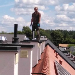 Gedbud Grzegorz Ged - Dekarz w brązowej koszulce polo i roboczych spodniach stoi na szczycie komina na tle błękitnego nieba z chmurami, widoczny fragment dachu z czerwoną dachówką i panelem słonecznym.