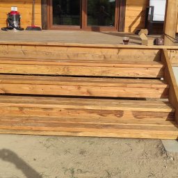 Gedbud Grzegorz Ged - Drewniane schody wejściowe do domu z tarasem, widoczny fragment elewacji, donica z krzewem i narzędzia na tarasie.