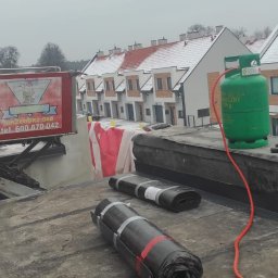 Gedbud Grzegorz Ged - Prace dekarskie na dachu budynku wielorodzinnego w trakcie realizacji, widoczne rolki papy, butla gazowa, narzędzia dekarskie oraz podnośnik z logo firmy.