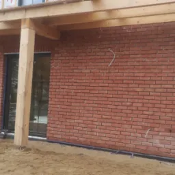 Murowana elewacja z czerwonej cegły klinkierowej w trakcie budowy, widoczne drewniane elementy konstrukcyjne dachu, okna z czarnymi ramami, oraz pracownik wykonujący prace wykończeniowe przy ścianie.