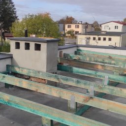 Gedbud Grzegorz Ged - Widok z góry na konstrukcję dachu z impregnowanych belek drewnianych, z kominem z otworami wentylacyjnymi, na tle zabudowy miejskiej w pochmurny dzień.