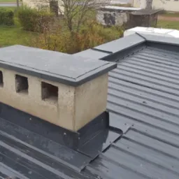 Szary blaszany dach z nową obróbką blacharską wokół komina z trzema otworami wentylacyjnymi, widok z góry na tle zieleni.