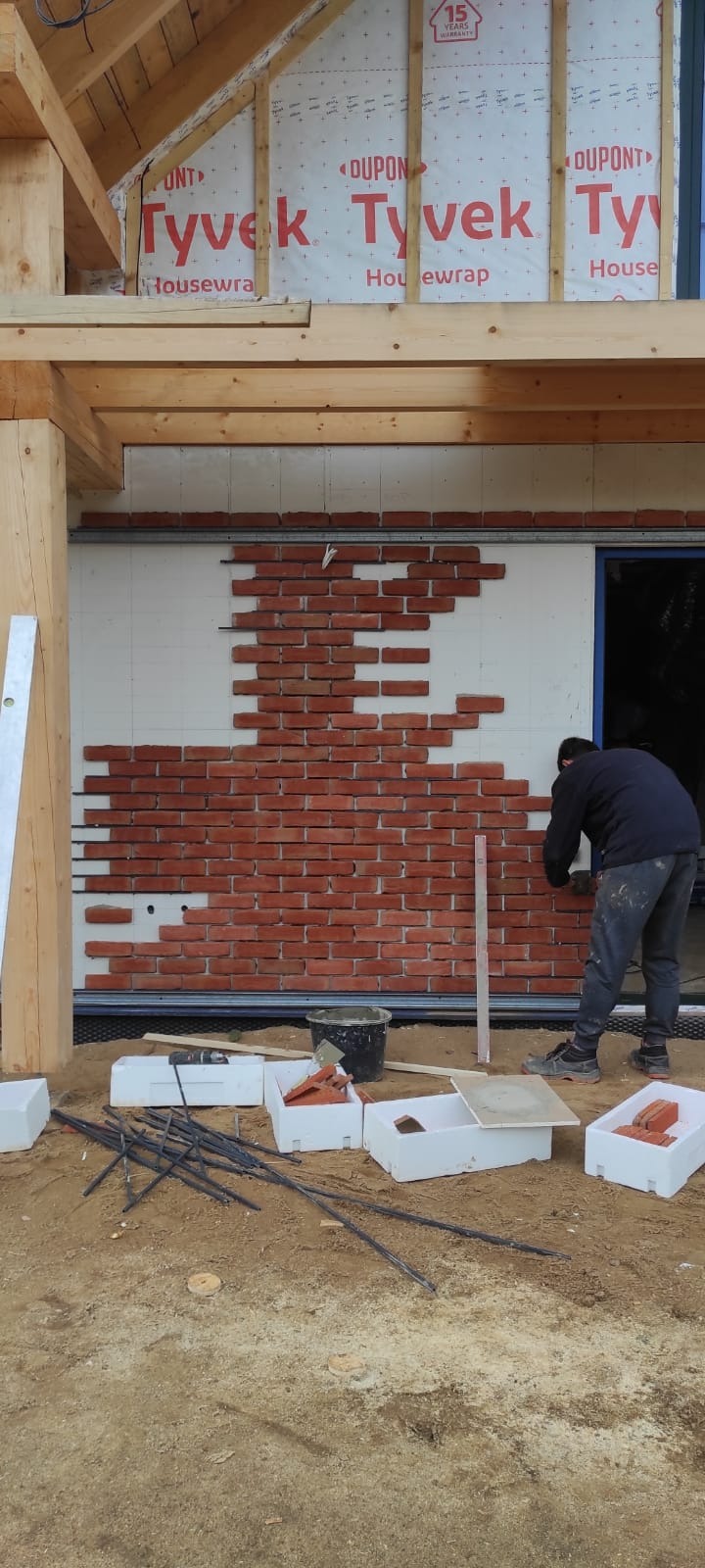 Mężczyzna w trakcie kładzenia cegieł klinkierowych na ocieplonej ścianie budynku, widoczne narzędzia, poziomica i przygotowane cegły w pojemnikach.