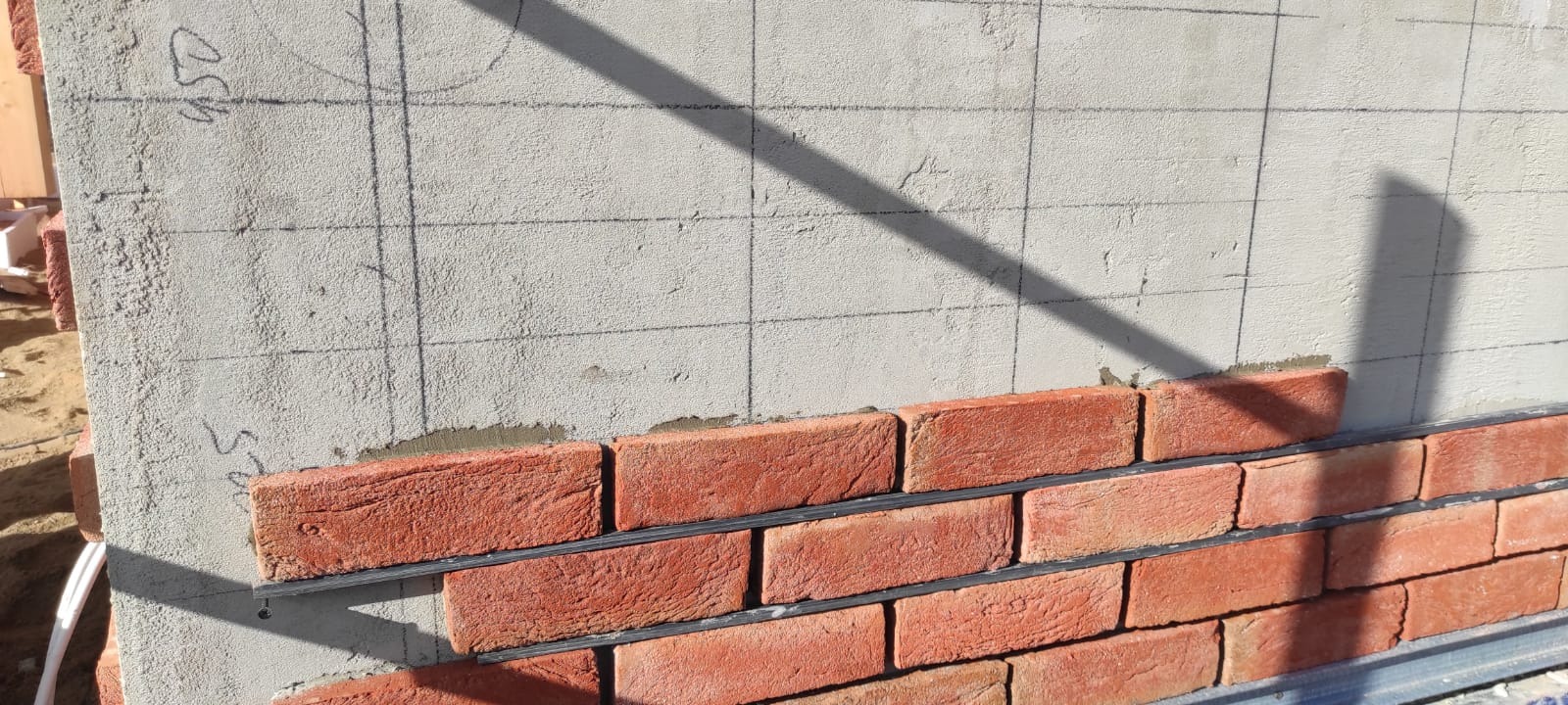 Montaż klinkieru na elewacji budynku z zaznaczoną siatką poziomowania, widoczny cień i fragment rusztowania.