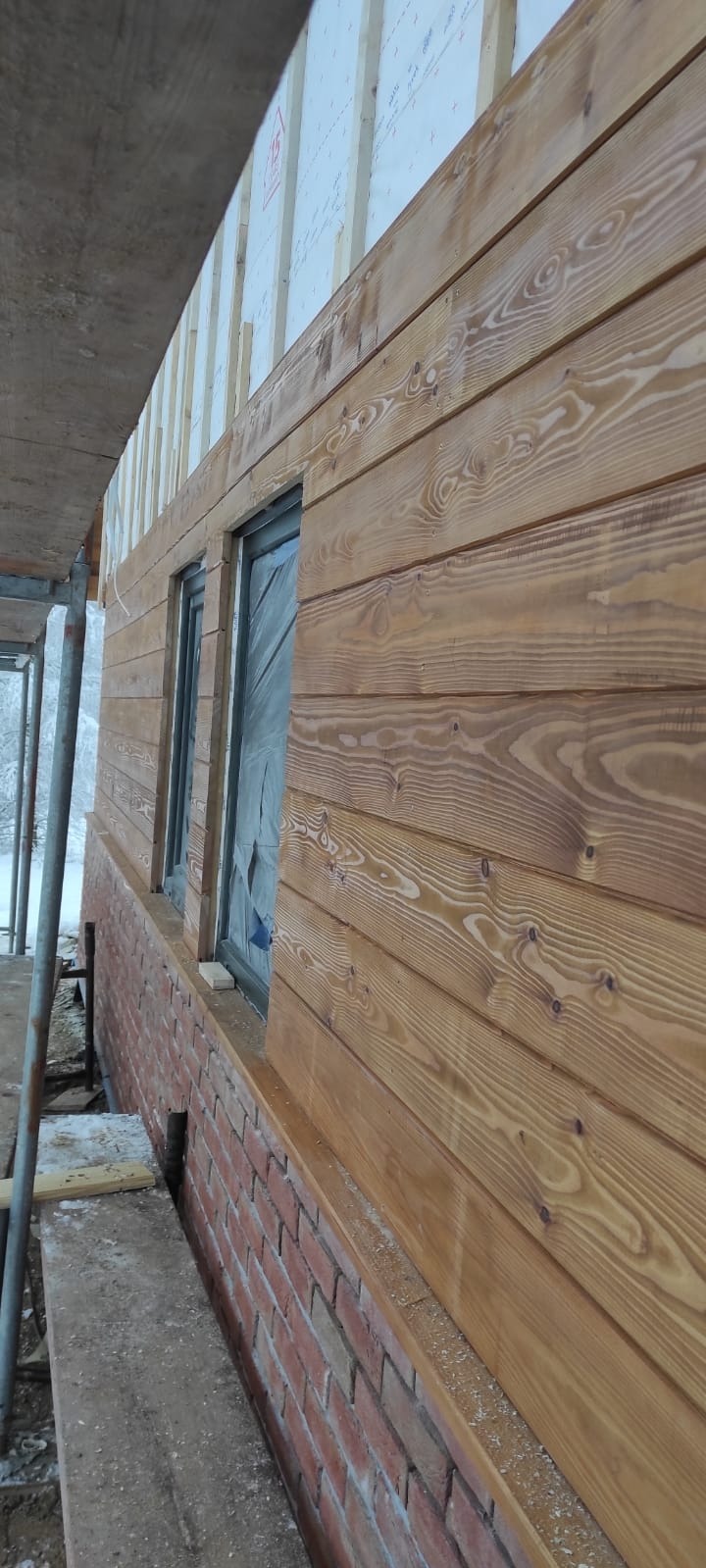 Ściana budynku z drewnianą elewacją w trakcie montażu, widoczne rusztowanie, okna zabezpieczone folią i ceglana podmurówka.