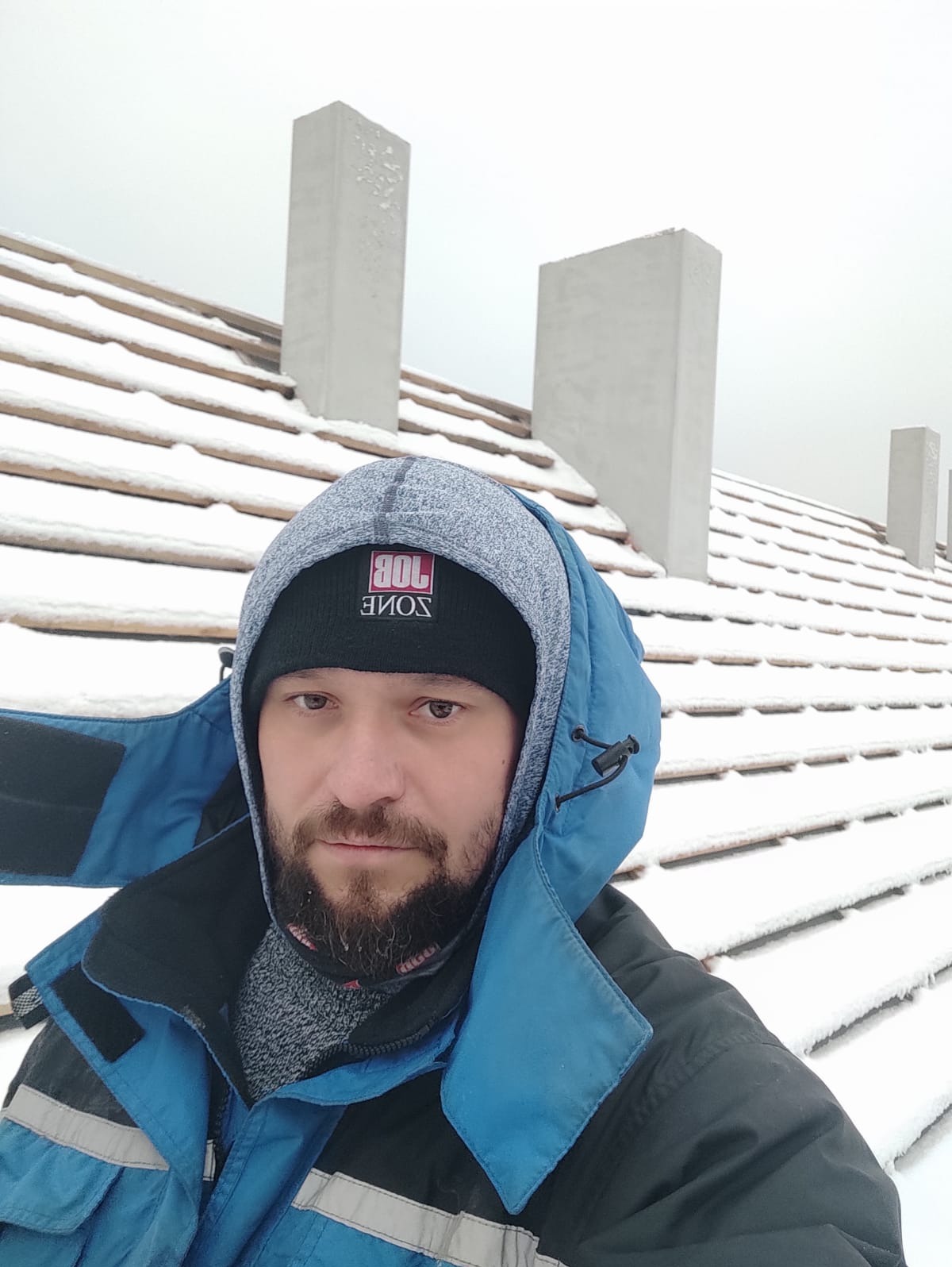 Selfie dekarza na tle zasypanego śniegiem dachu z kominami, ubrany w zimową kurtkę i czapkę z logo.