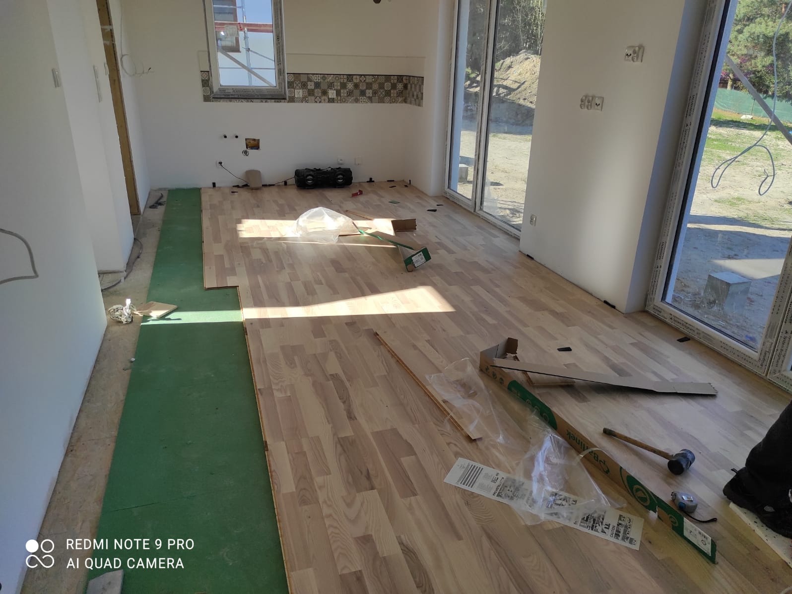 Wnętrze w trakcie układania drewnianej podłogi, widoczne panele, podkład z zielonej pianki, narzędzia i opakowania po panelach, duże okna z widokiem na zewnątrz.