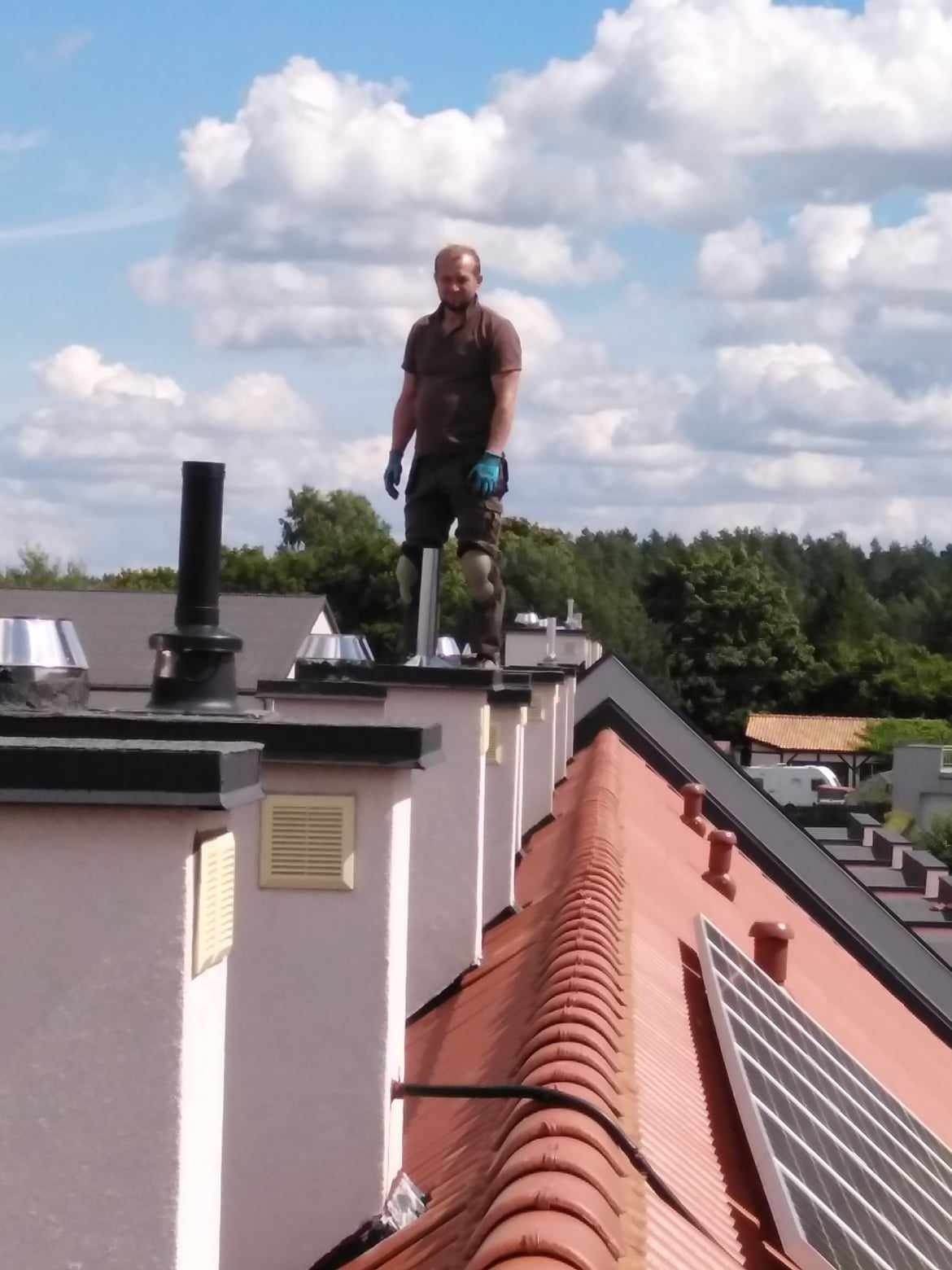 Dekarz w brązowej koszulce polo i roboczych spodniach stoi na szczycie komina na tle błękitnego nieba z chmurami, widoczny fragment dachu z czerwoną dachówką i panelem słonecznym.