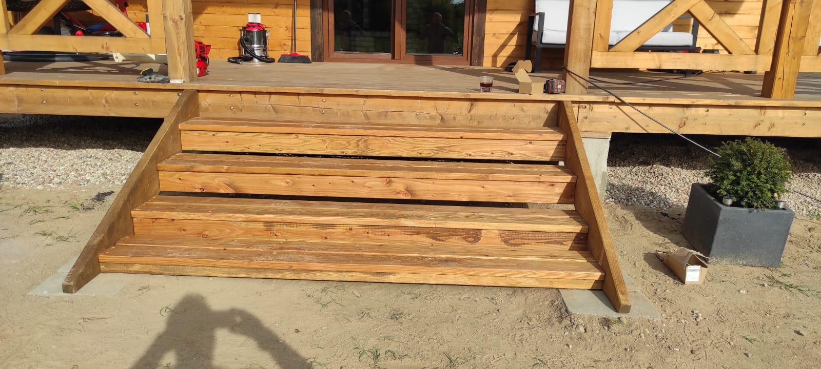 Drewniane schody wejściowe do domu z tarasem, widoczny fragment elewacji, donica z krzewem i narzędzia na tarasie.