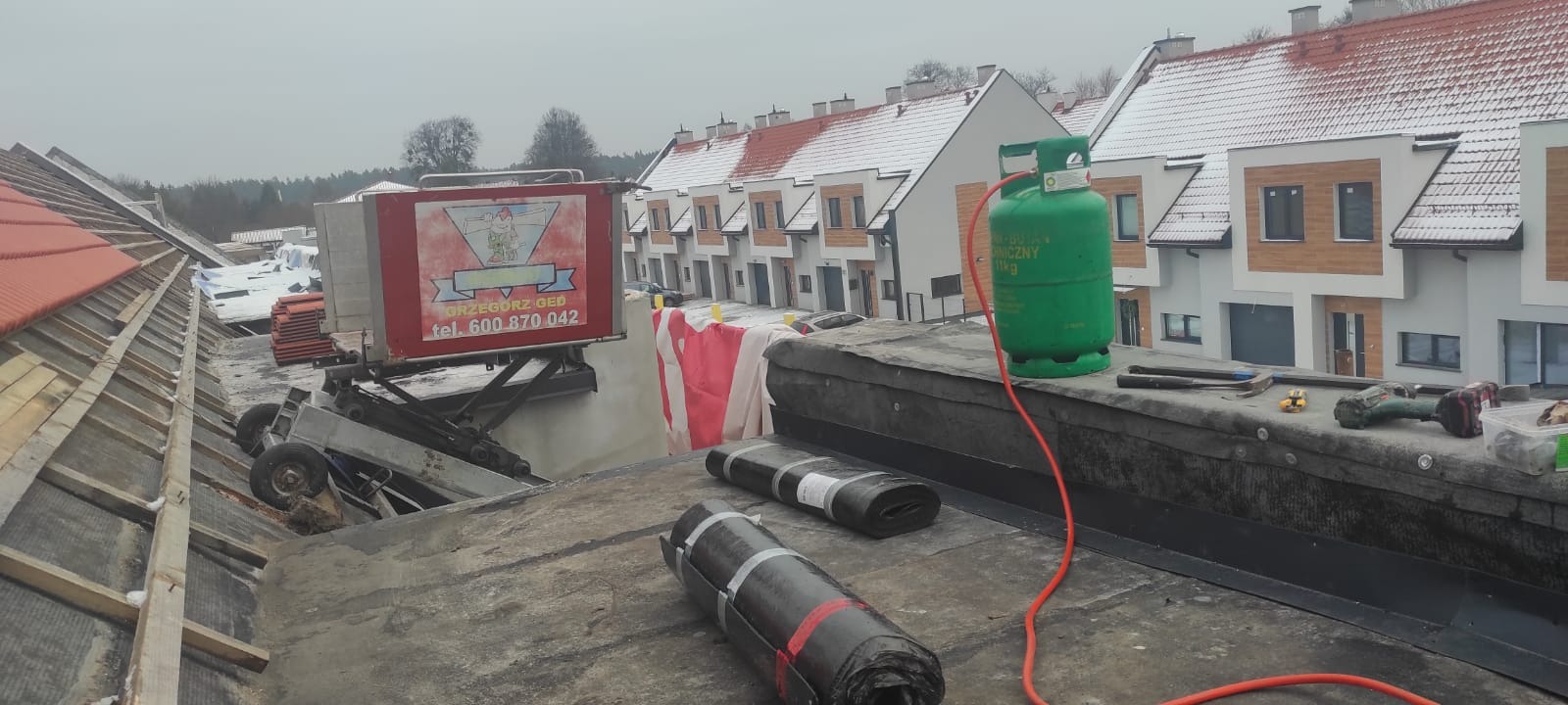 Prace dekarskie na dachu budynku wielorodzinnego w trakcie realizacji, widoczne rolki papy, butla gazowa, narzędzia dekarskie oraz podnośnik z logo firmy.