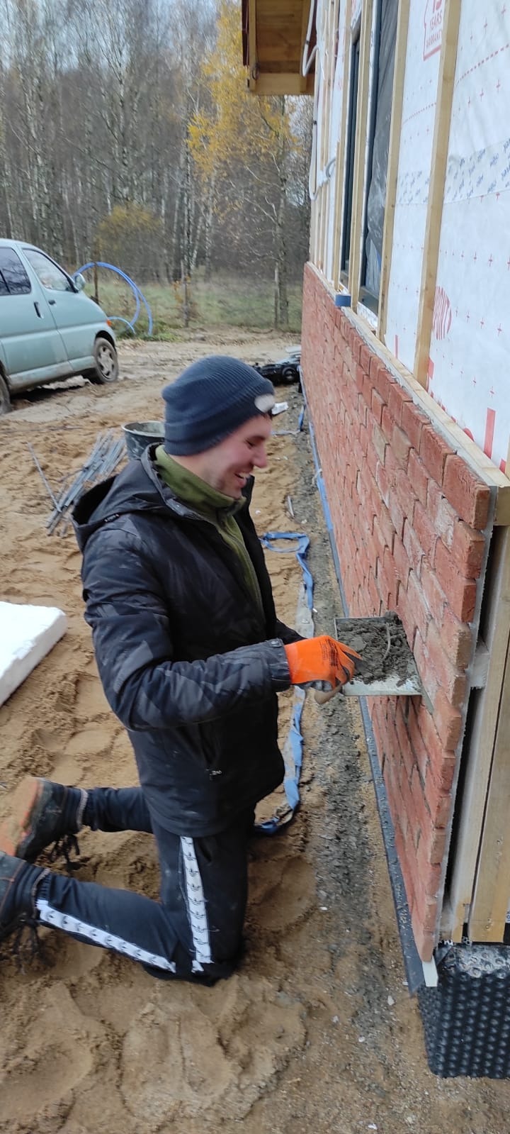 Uśmiechnięty murarz w czapce i pomarańczowych rękawicach, klęczący na piasku, układa cegły na elewacji budynku w trakcie budowy.