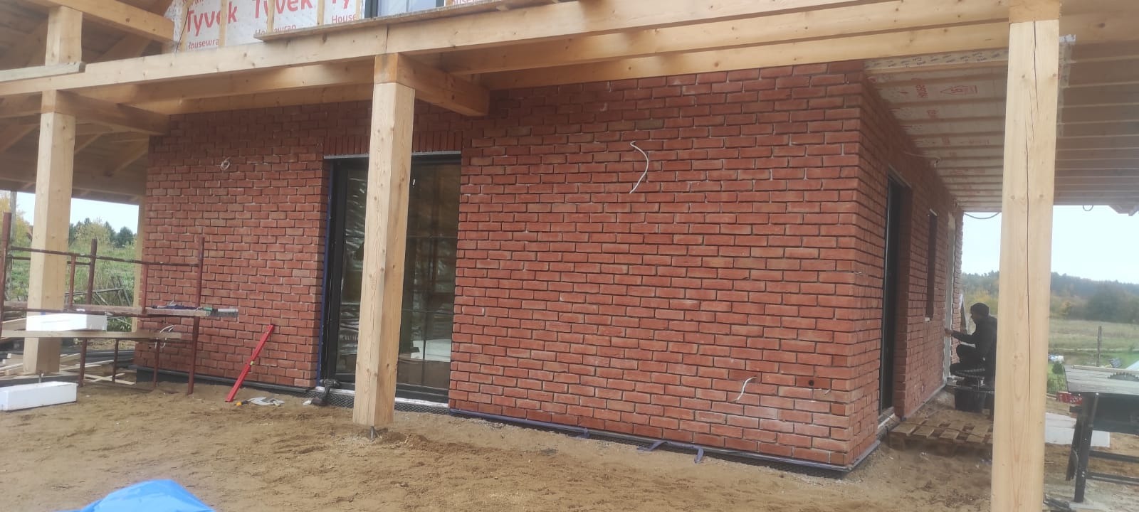 Murowana elewacja z czerwonej cegły klinkierowej w trakcie budowy, widoczne drewniane elementy konstrukcyjne dachu, okna z czarnymi ramami, oraz pracownik wykonujący prace wykończeniowe przy ścianie.