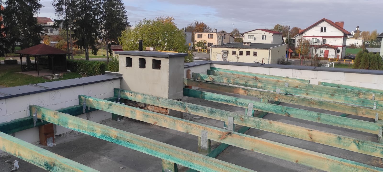 Widok z góry na konstrukcję dachu z impregnowanych belek drewnianych, z kominem z otworami wentylacyjnymi, na tle zabudowy miejskiej w pochmurny dzień.