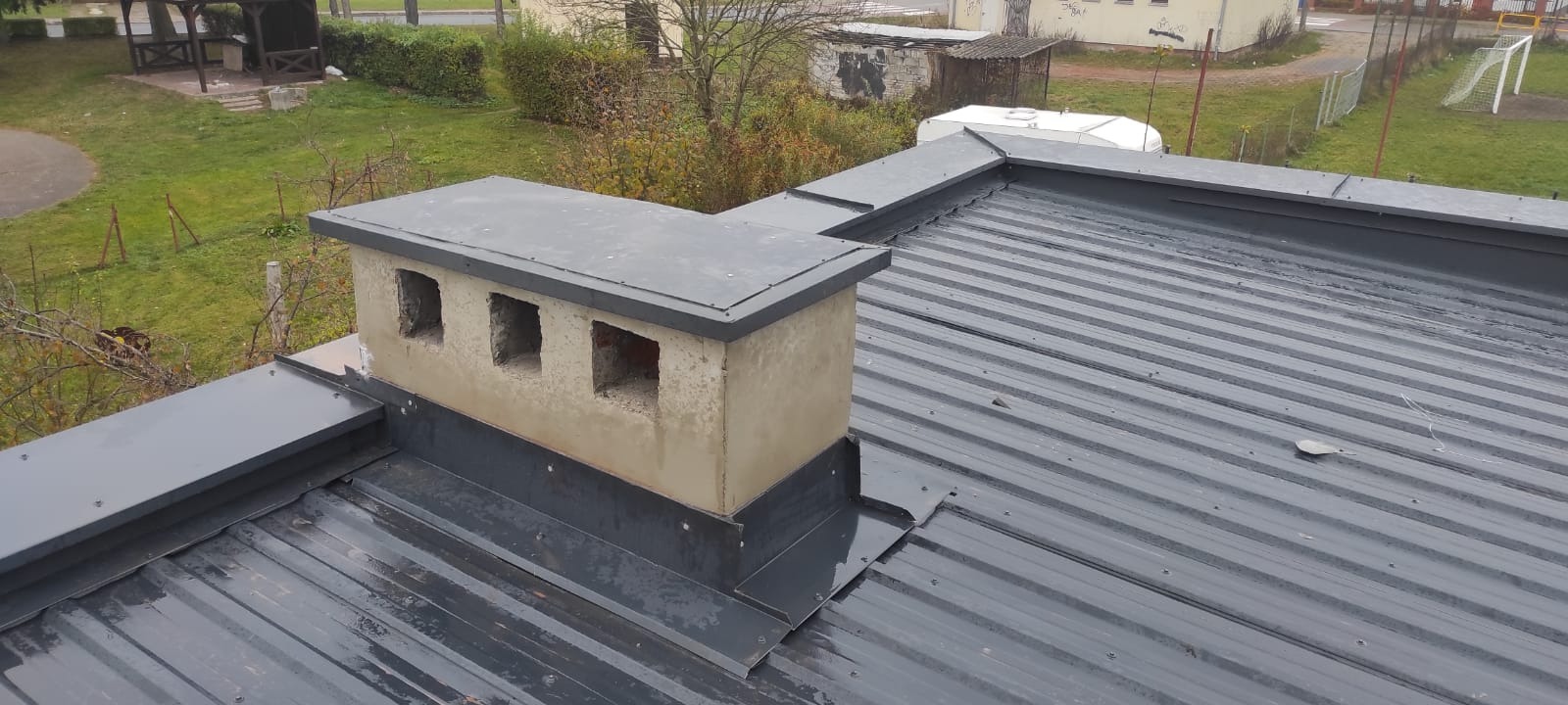 Szary blaszany dach z nową obróbką blacharską wokół komina z trzema otworami wentylacyjnymi, widok z góry na tle zieleni.