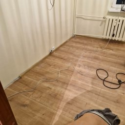 Osoba prywatna - Fragment pokoju w trakcie prac wykończeniowych, widoczne panele podłogowe, listwy przypodłogowe, grzejnik, gniazdko elektryczne oraz przewody na podłodze.