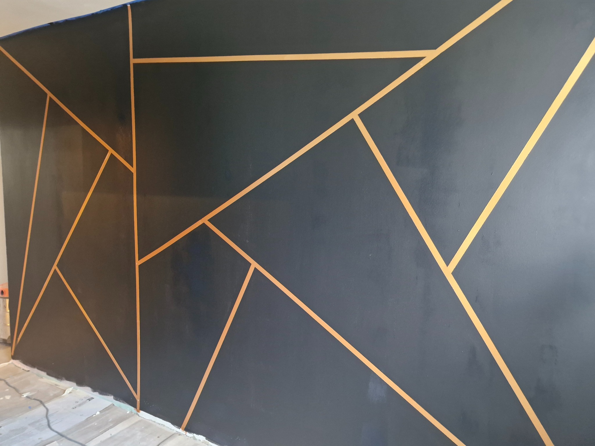 Ściana pomalowana na ciemny grafit z geometrycznym wzorem wykonanym złotą taśmą malarską, widoczne niedoskonałości przy podłodze.
