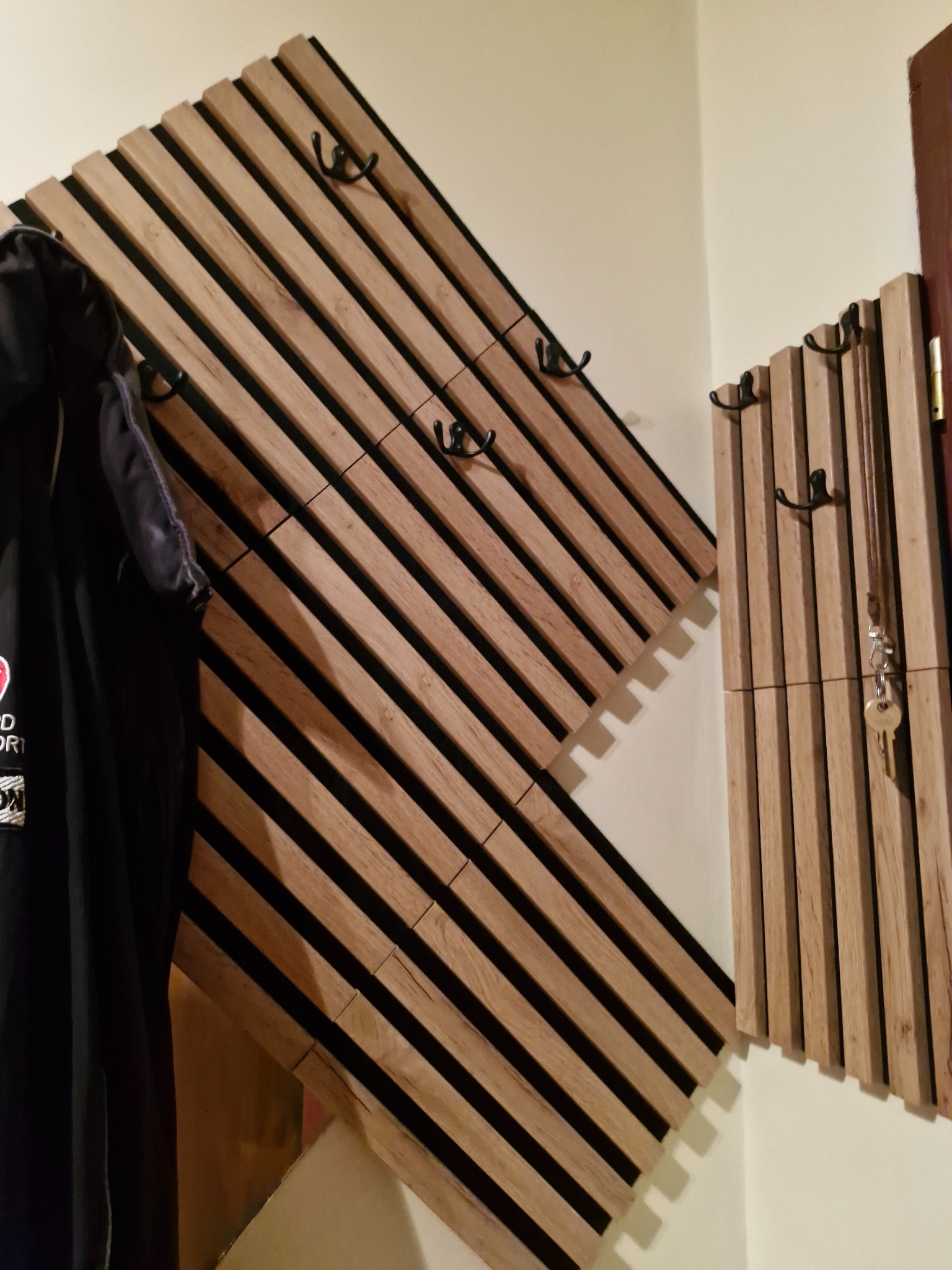 Drewniany wieszak panelowy z czarnymi akcentami i haczykami, jeden z wieszaków w kształcie rombu, drugi pionowy, na jednym wisi czarna kurtka, na drugim klucze