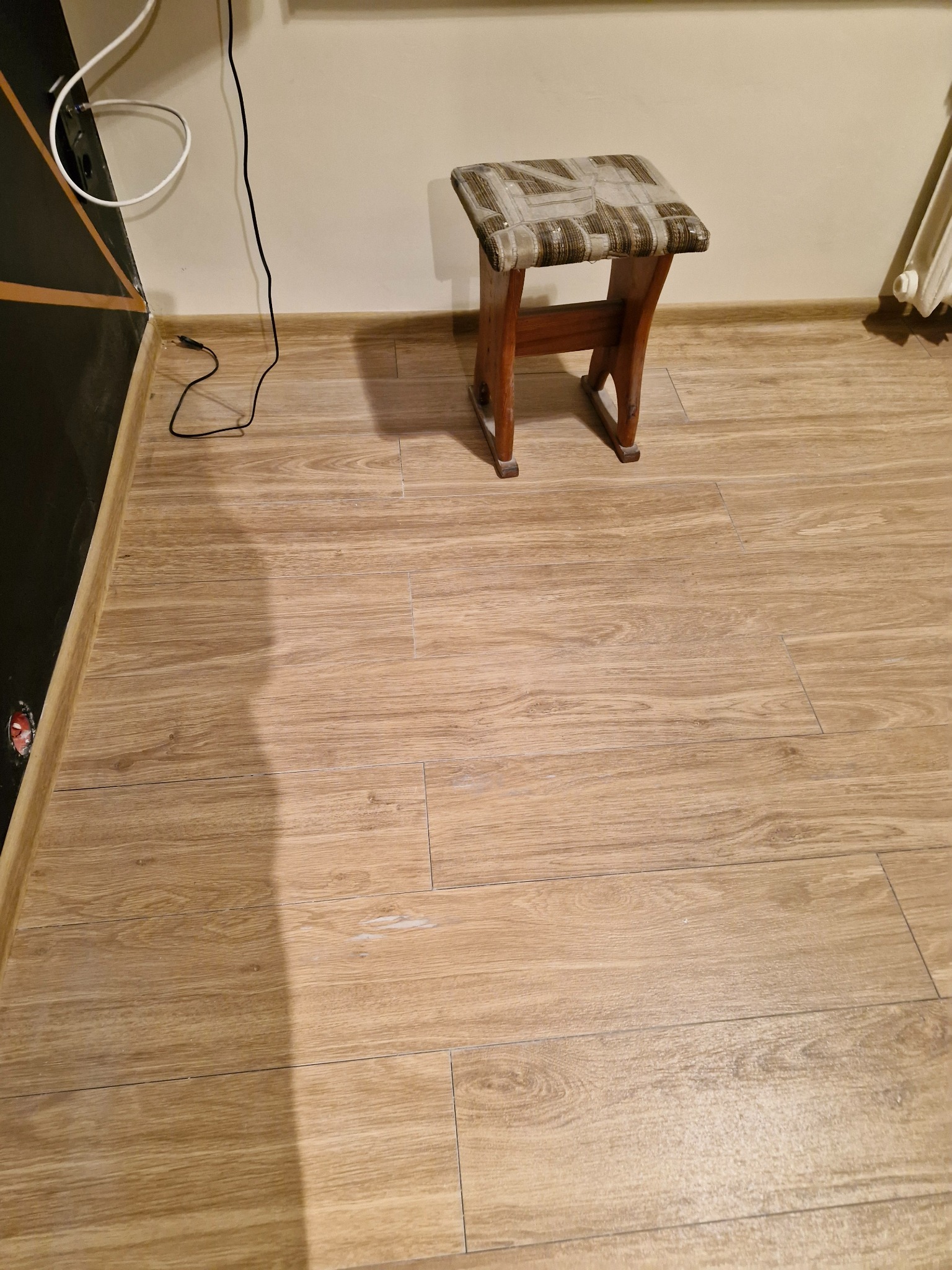 Ułożona podłoga z paneli imitujących drewno, widoczny fragment ściany, grzejnik i taboret z drewnianymi nogami.