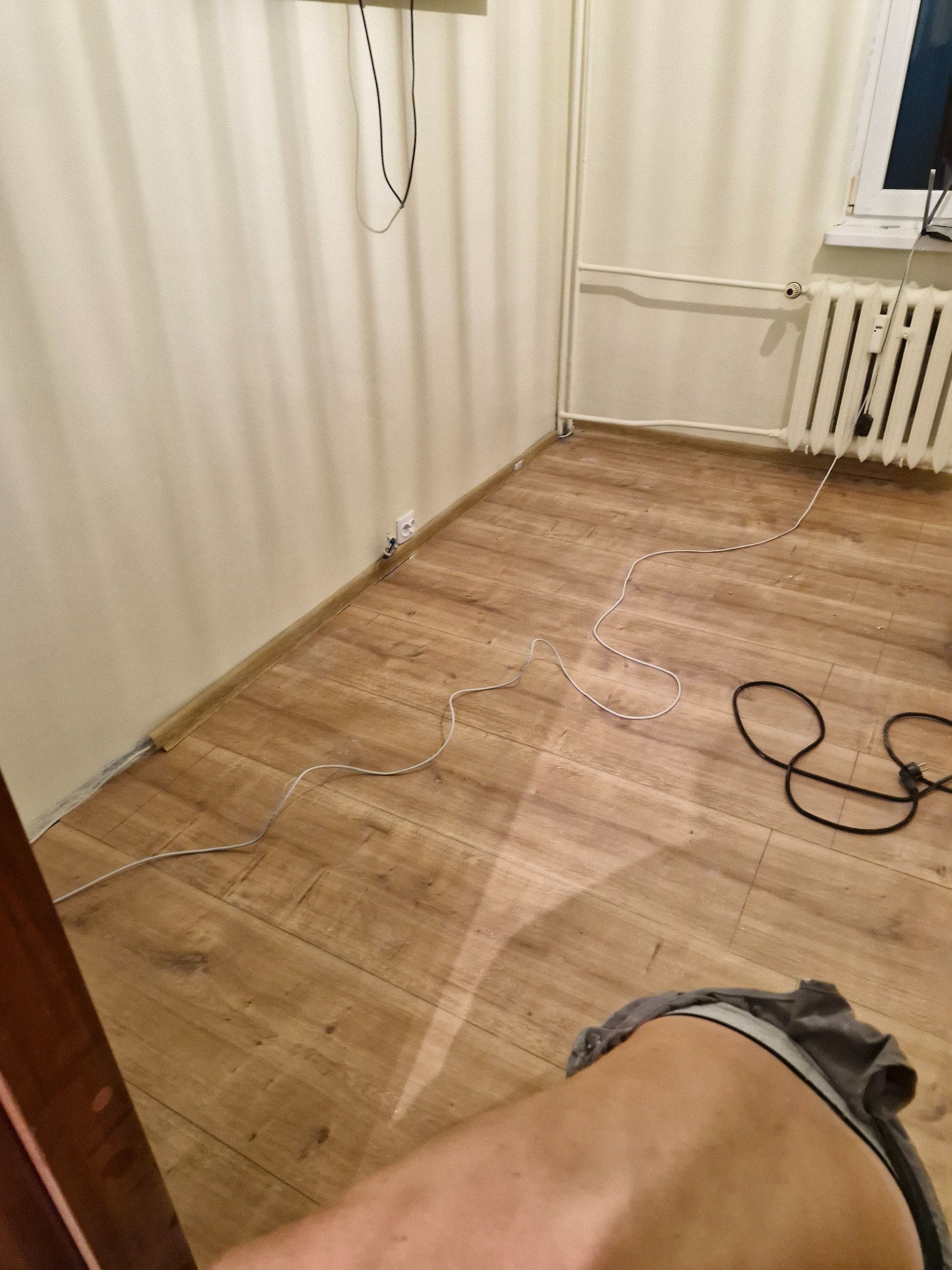 Fragment pokoju w trakcie prac wykończeniowych, widoczne panele podłogowe, listwy przypodłogowe, grzejnik, gniazdko elektryczne oraz przewody na podłodze.