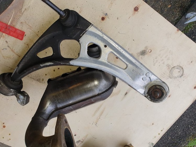 Metalowe wahacze samochodowe, jeden pomalowany na srebrno w połowie, drugi czarny, leżące obok zardzewiałego katalizatora na drewnianym podłożu.