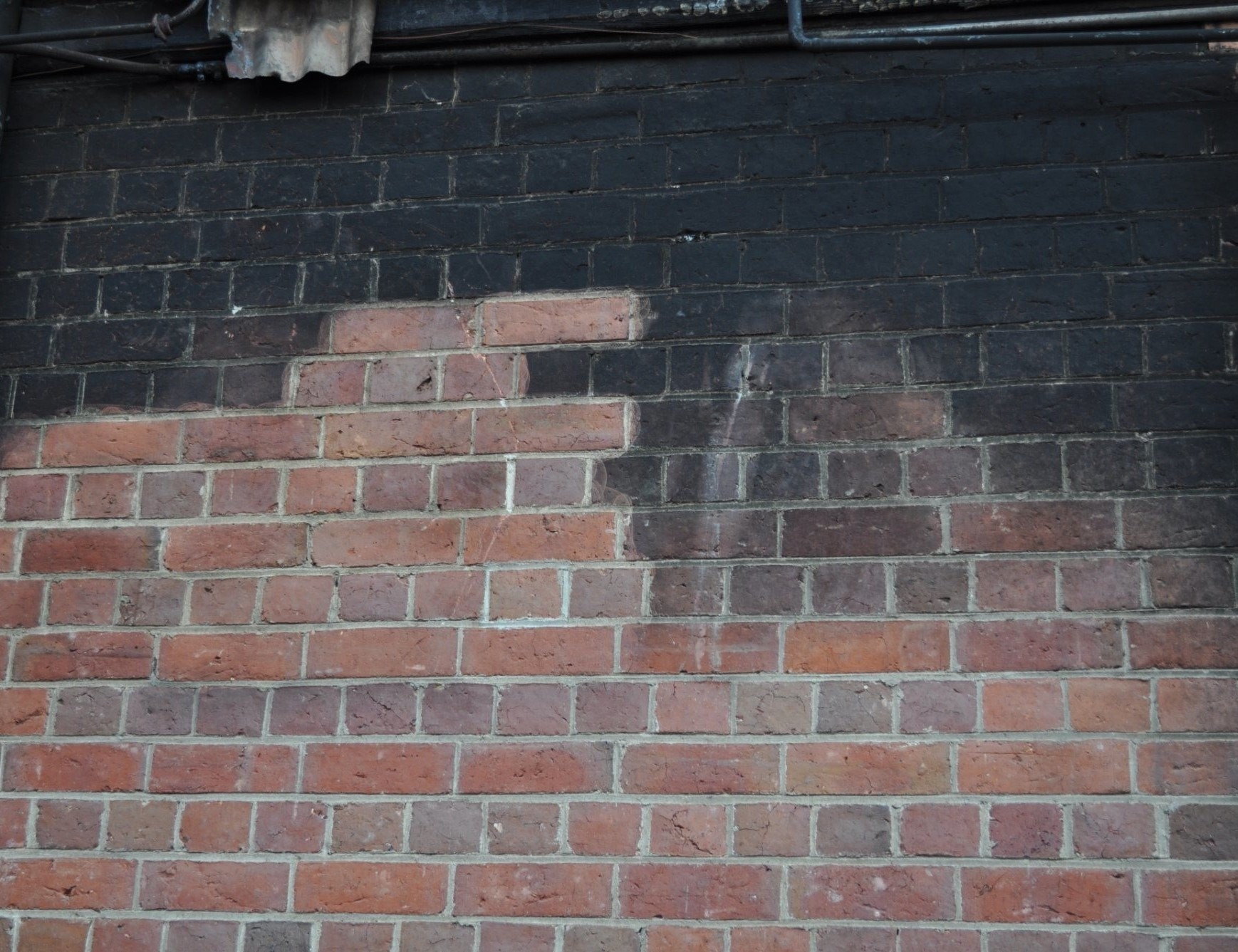 Częściowo oczyszczona ceglana ściana z widocznym kontrastem między czystą, czerwoną cegłą a zaciemnioną, brudną górną częścią. Widoczne ślady spływającej wody po czyszczeniu.