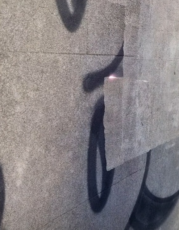 Szara, betonowa ściana z fragmentami czarnego graffiti i odspojonym fragmentem tynku, ukazująca surową fakturę i ślady upływu czasu.