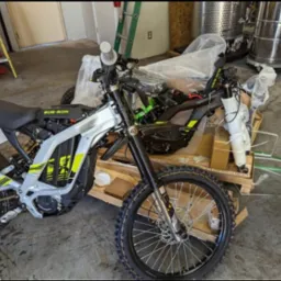 Nowy, częściowo zmontowany elektryczny motocykl terenowy Sur-Ron, z elementami owiniętymi folią, stoi na drewnianej palecie w magazynie; w tle drabina i stalowy zbiornik.