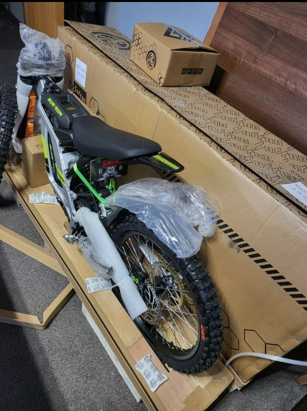 Nowy elektryczny motocykl crossowy Sur-Ron zapakowany w kartonowe pudełko z widocznymi oponami terenowymi, błotnikiem owiniętym folią i elementami ramy zabezpieczonymi pianką.