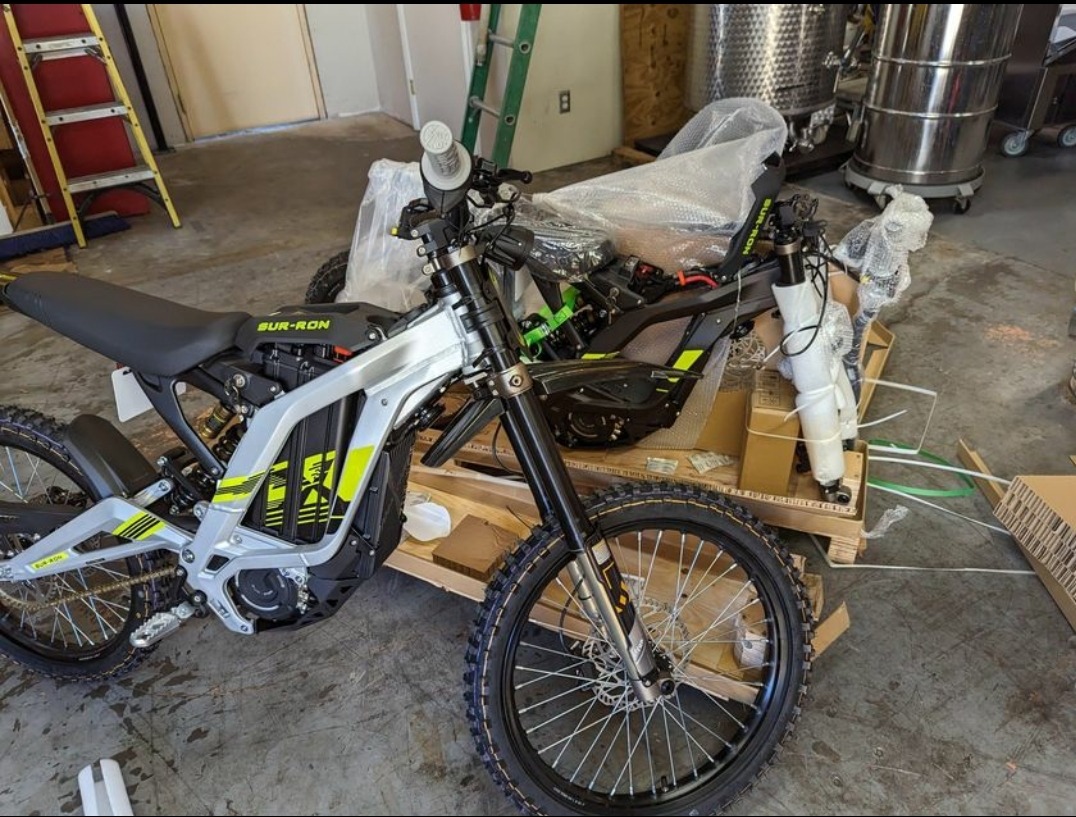 Nowy, częściowo zmontowany elektryczny motocykl terenowy Sur-Ron, z elementami owiniętymi folią, stoi na drewnianej palecie w magazynie; w tle drabina i stalowy zbiornik.