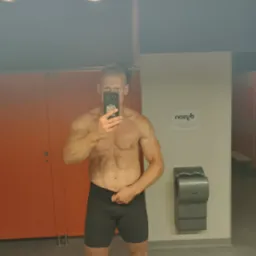 Mężczyzna z wyrzeźbioną sylwetką, robiący selfie w szatni siłowni, widoczny automat do suszenia rąk Dyson i pomarańczowe drzwi do toalet.