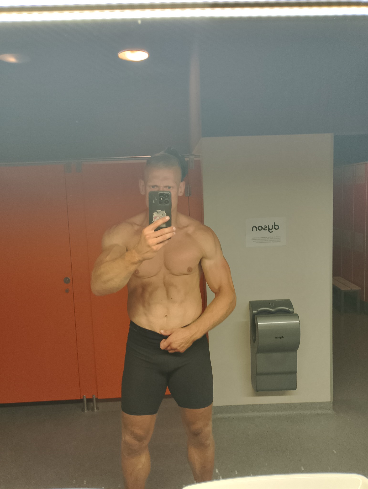 Mężczyzna z wyrzeźbioną sylwetką, robiący selfie w szatni siłowni, widoczny automat do suszenia rąk Dyson i pomarańczowe drzwi do toalet.