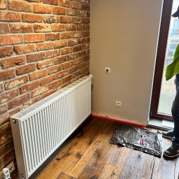 Biały grzejnik na tle ceglanej ściany w Środzie Śląskiej. Widoczne narzędzia i przygotowane miejsce do montażu. Podłoga drewniana, deski z wyraźnym usłojeniem.