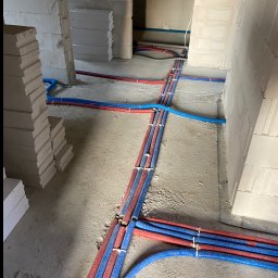 Nowa instalacja wodno-kanalizacyjna i CO w surowym budynku. Czerwone i niebieskie rury prowadzone po podłodze, przymocowane opaskami. Widoczne białe bloczki.