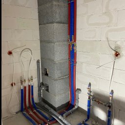 Instalacja wodna z rur PEX w kolorach niebieskim i czerwonym, z mosiężnymi złączkami, zamocowana do ściany z betonu komórkowego i szarego komina wentylacyjnego.