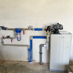 Renova - Starannie wykonana instalacja hydrauliczna z niebieskim filtrem wody, wodomierzem i dużym zmiękczaczem, zamontowana na jasnej, niewykończonej ścianie.