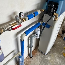 Renova - Nowoczesna instalacja hydrauliczna z niebieskimi rurami izolacyjnymi, manometrem i filtrem wody, zamontowana na jasnej ścianie, z widocznym zaworem PERFEKT SYSTEM.