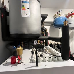Renova - Kompletna instalacja grzewcza z szarym bojlerem 93L, izolowanymi rurami, dwoma naczyniami wzbiorczymi (niebieskim i białym) i panelem sterującym z okablowaniem.