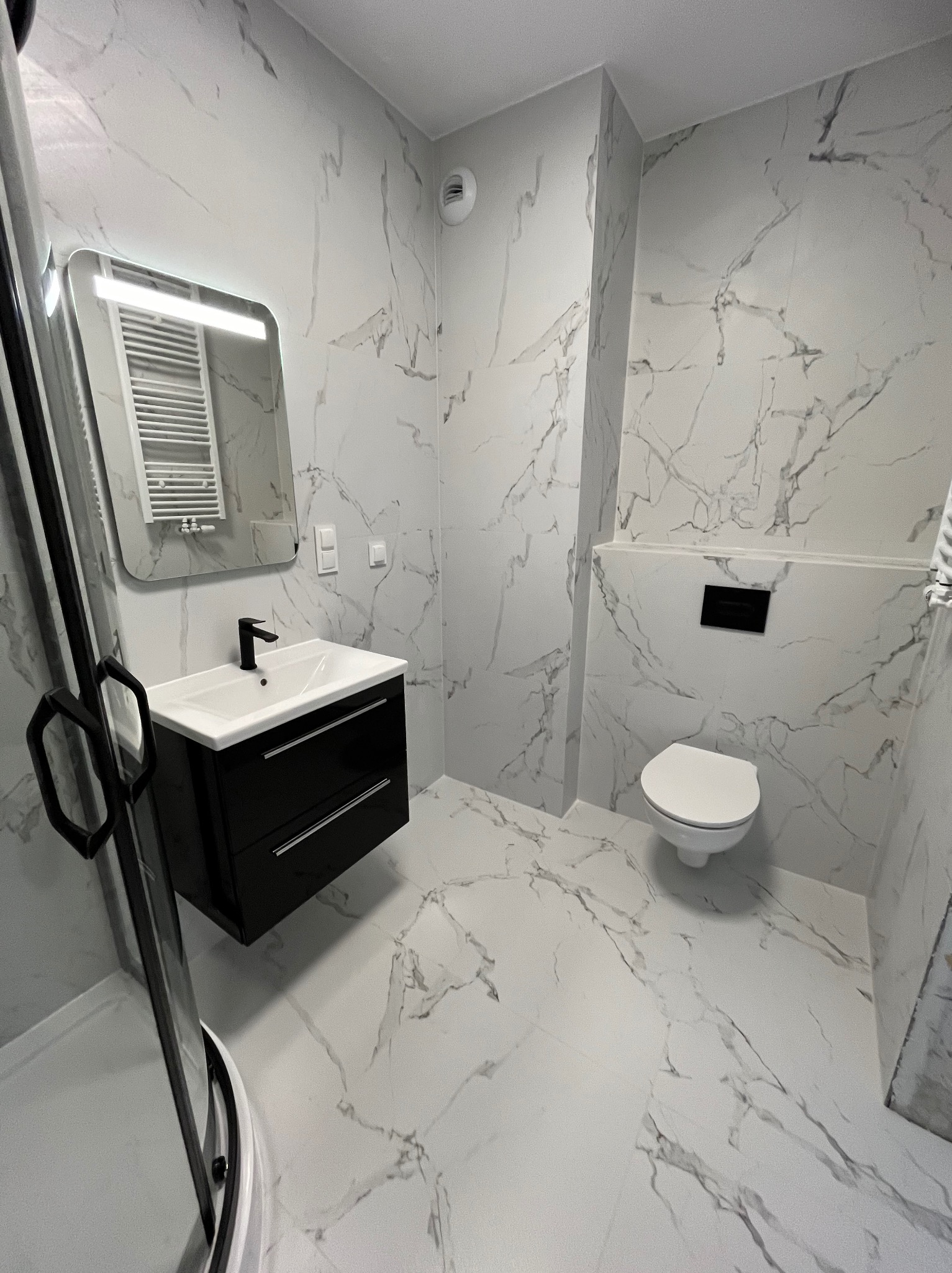 Wykończona łazienka w bieli i czerni: marmurowe płytki, podwieszana szafka z umywalką, toaleta podtynkowa i kabina prysznicowa z czarnymi detalami.
