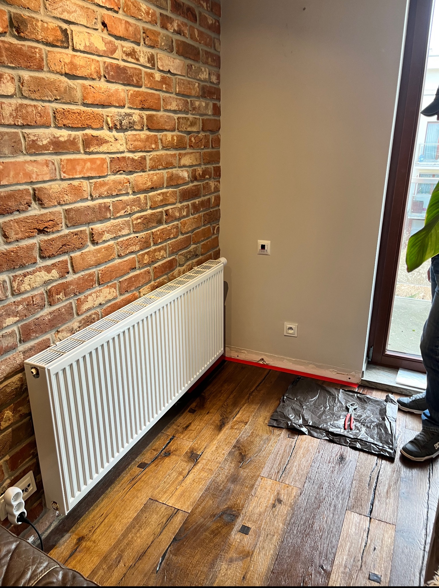 Biały grzejnik na tle ceglanej ściany w Środzie Śląskiej. Widoczne narzędzia i przygotowane miejsce do montażu. Podłoga drewniana, deski z wyraźnym usłojeniem.