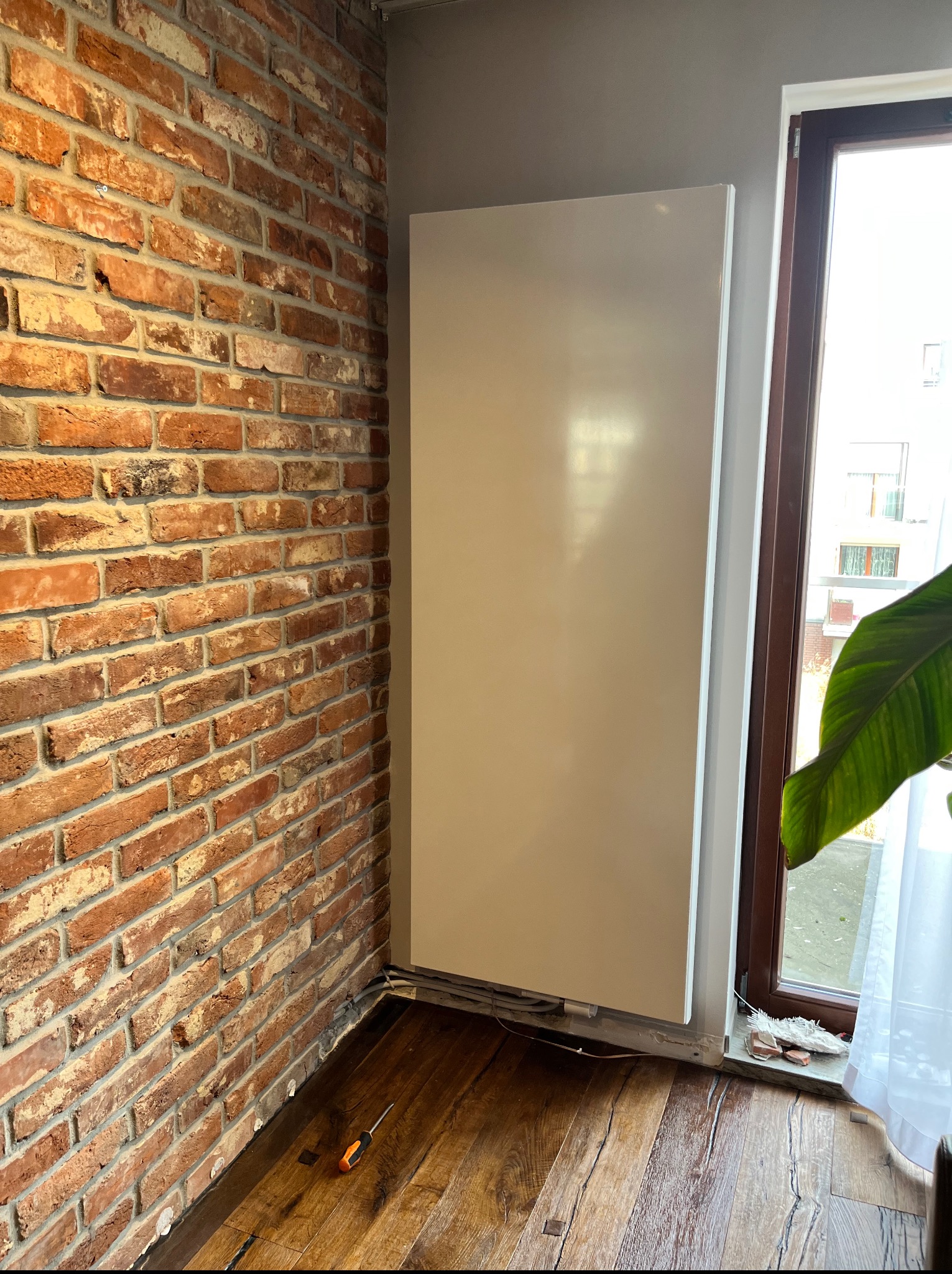 Biały, płaski grzejnik na tle ceglanej ściany i okna. Widoczne drewniane panele podłogowe i narzędzie. Nietypowe połączenie stylów w wykończeniu wnętrza.